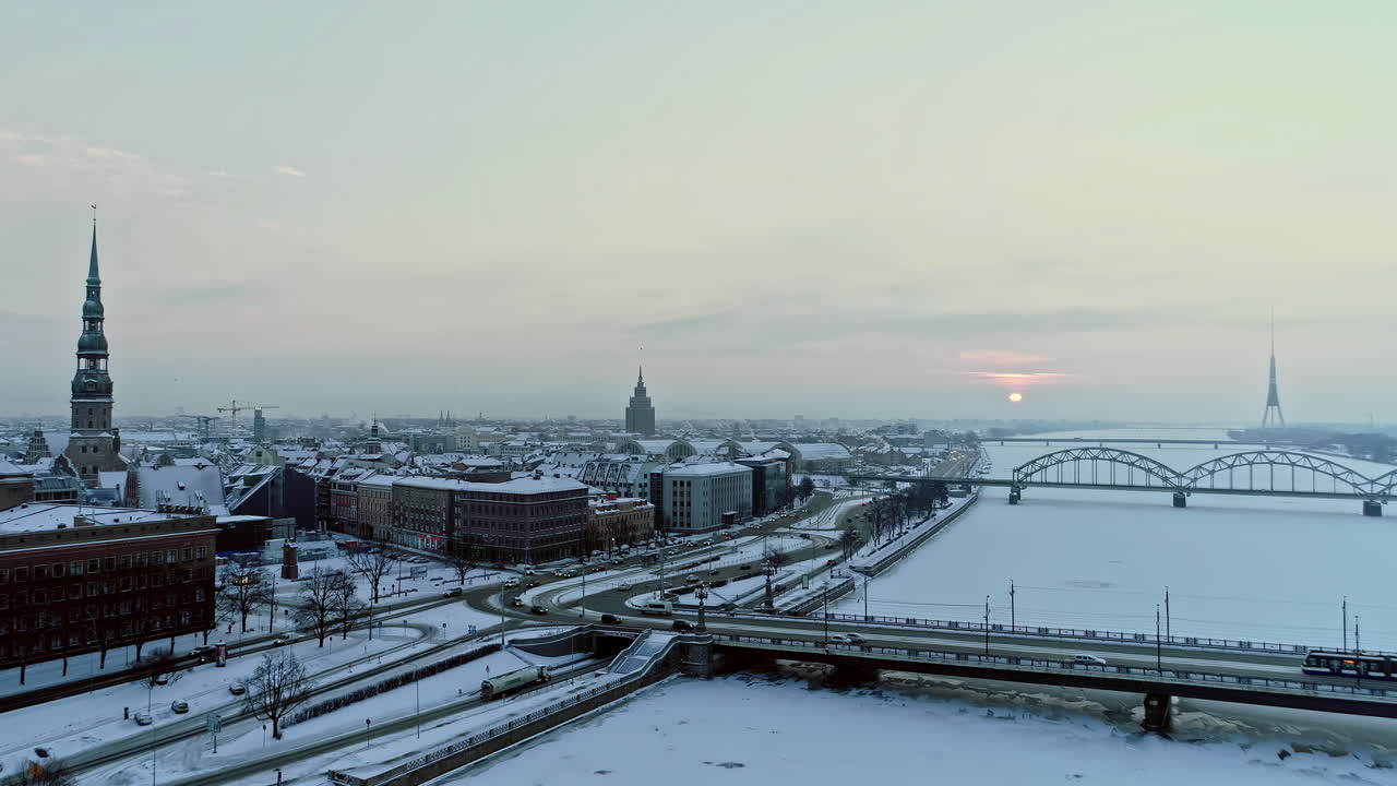 toma aérea hacia atrás de la ciudad de riga durante el día de invierno nevado en la mañana - río daugava congelado, conducción de automóviles, puente y catedral durante el amanecer - riga capital de letonia