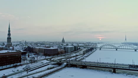 aerial backwards shot of riga city during snowy winter day in the morning - frozen daugava river, driving cars,bridge and cathedral during sunrise - riga capital of latvia