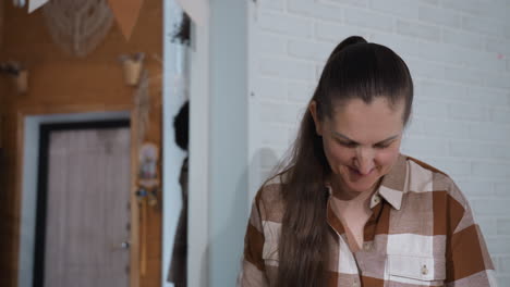 woman wearing plaid shirt and long ponytail smiling gently while looking down and moving hands gracefully in bright modern kitchen with brick wall background capturing warm candid moment