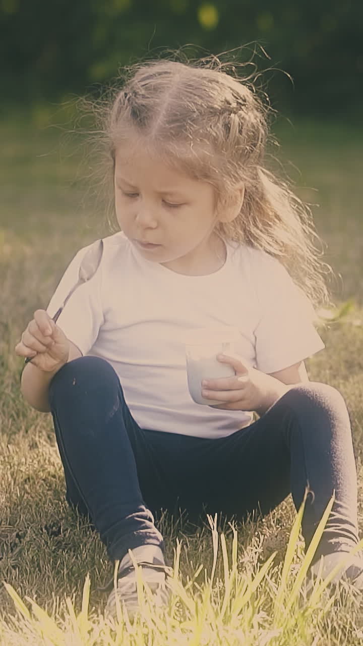 little girl with long fair hair sits on grass looks at large metal spoon holding white yogurt container slow motion