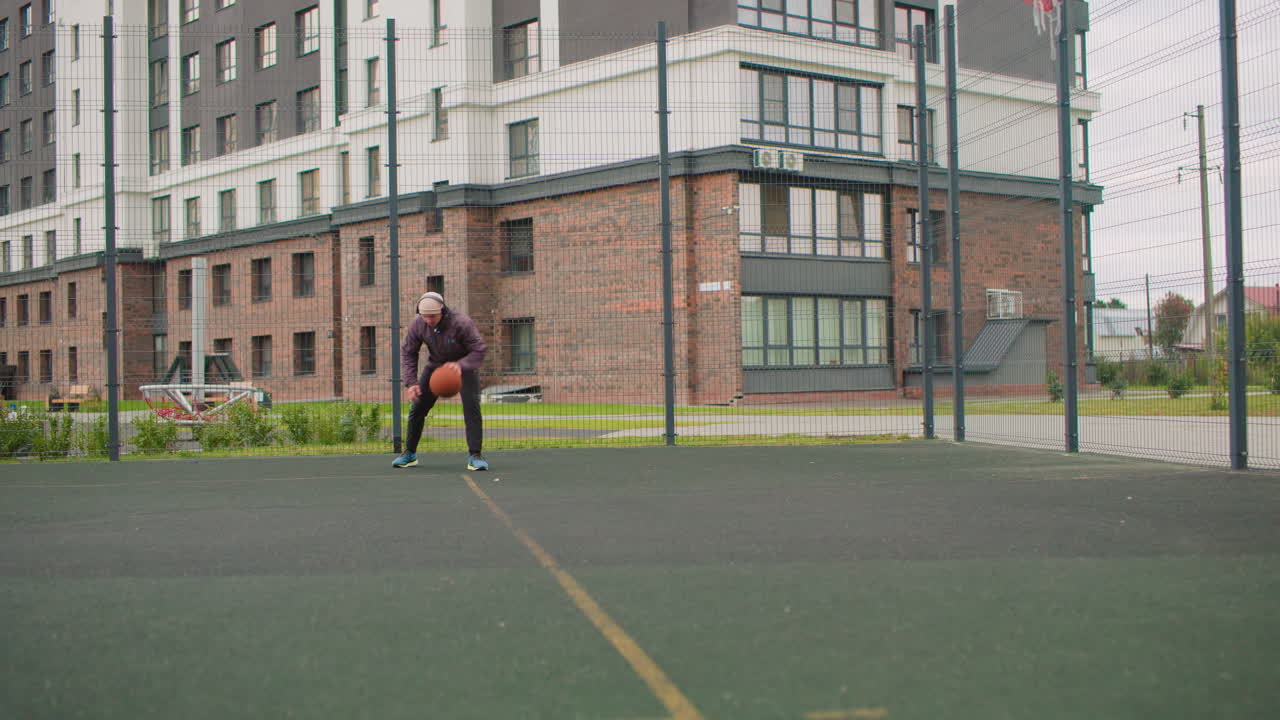 Un hombre mayor practica ejercicios en solitario en una cancha de baloncesto con edificios de apartamentos al fondo; camina hacia la canasta, bota la pelota, lanza y recoge el rebote. Cielo nublado, valla metálica y superficie de asfalto.