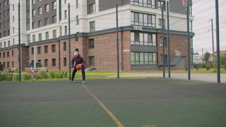 Uomo anziano su un campo da basket che si esercita da solo in palleggio sullo sfondo di un edificio residenziale, cammina verso il canestro, palleggia, tira, recupera la palla, cielo nuvoloso, recinzione metallica, superficie asfaltata