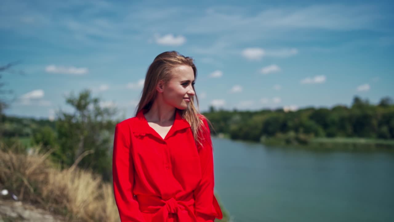 Pretty woman in summer outdoors. Beautiful girl with long hair on the river background. Young model in red dress touching her hair and smiling. Slow motion.