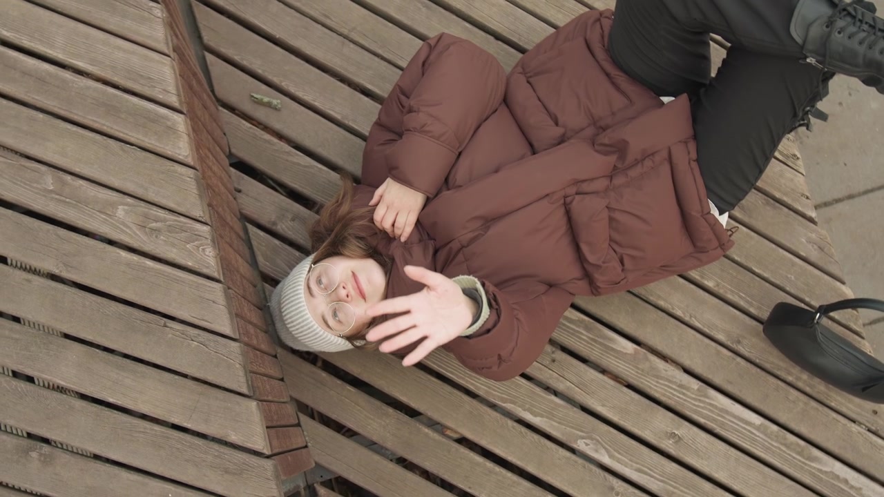 Smilende kvinde på træplanker i brun frakke og strikhue, vinker og peger med livligt udtryk, lille taske ved siden, luftfoto fanger diagonale teksturer og glad stemning.