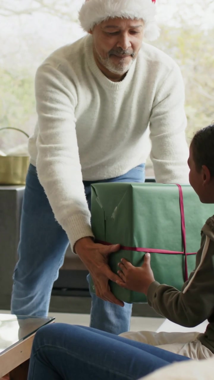 Video verticale: Un uomo con cappello di Babbo Natale e maglione bianco entra in casa e dà una scatola verde a un bambino.