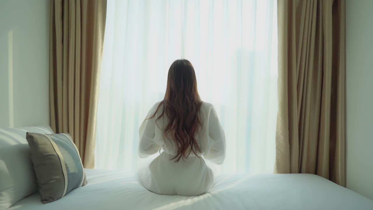 Elegant brunette in white bathrobe sits on French bed and stretches, behind