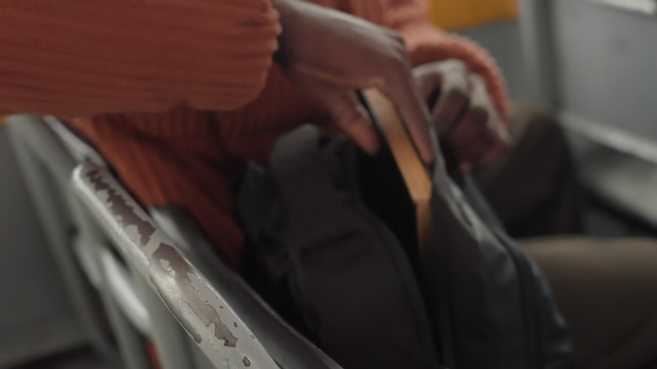 Viajero negro en autobús sacando un libro, manos con suéter naranja abriendo la mochila, asiento de metal, primer plano del movimiento y la textura, ambiente tranquilo y reflexivo, roles de estudiante, periodista, investigador acomodándose.
