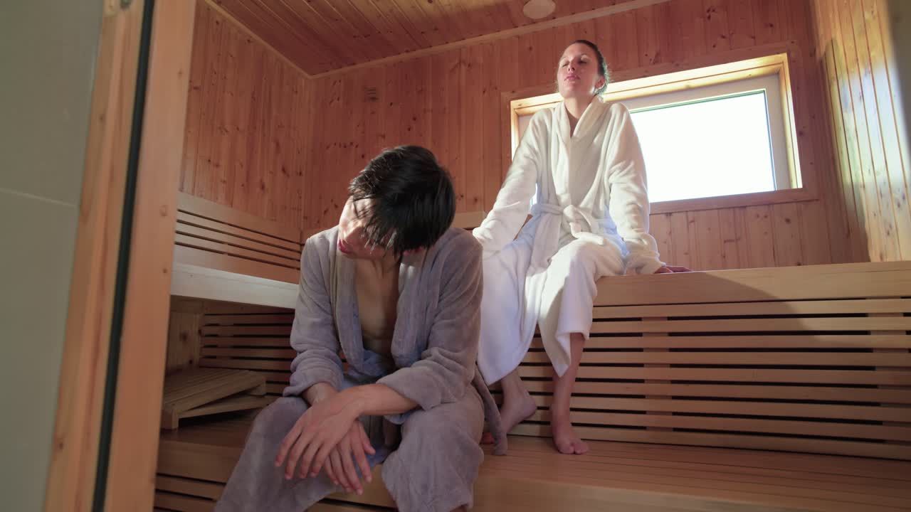 Coppia in accappatoio seduta su panche di legno della sauna, che respira profondamente e reagisce al calore della sauna.