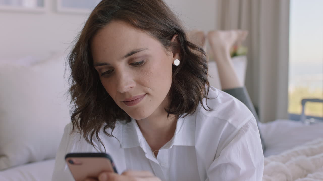 스마트폰을 사용하는 아름다운 여성 침대에서 휴식을 취하고 문자 메시지를 보내고 소셜 미디어 메시지를 공유하고 온라인 쇼핑을 즐기고 있습니다.