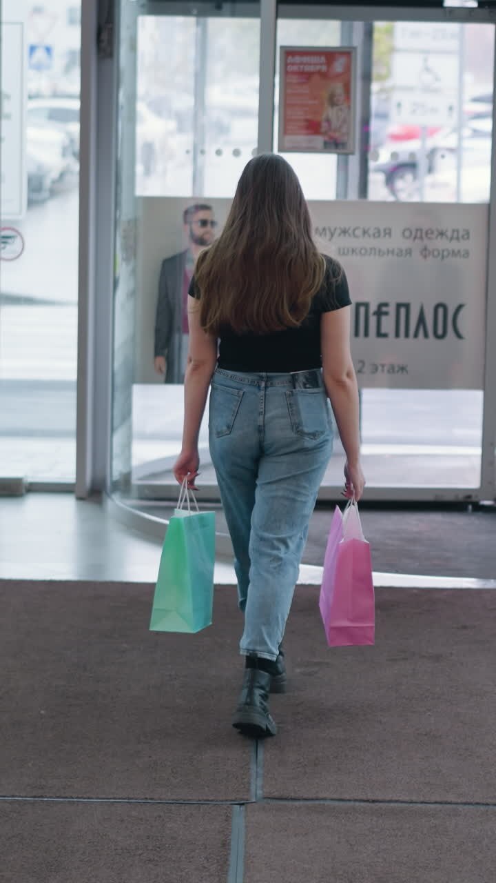 Donna con borse della spesa che cammina verso l'uscita in un centro commerciale moderno, persona che passa, porte girevoli in movimento, abbigliamento alla moda, stivali casual, esperienza di shopping urbana con illuminazione intensa