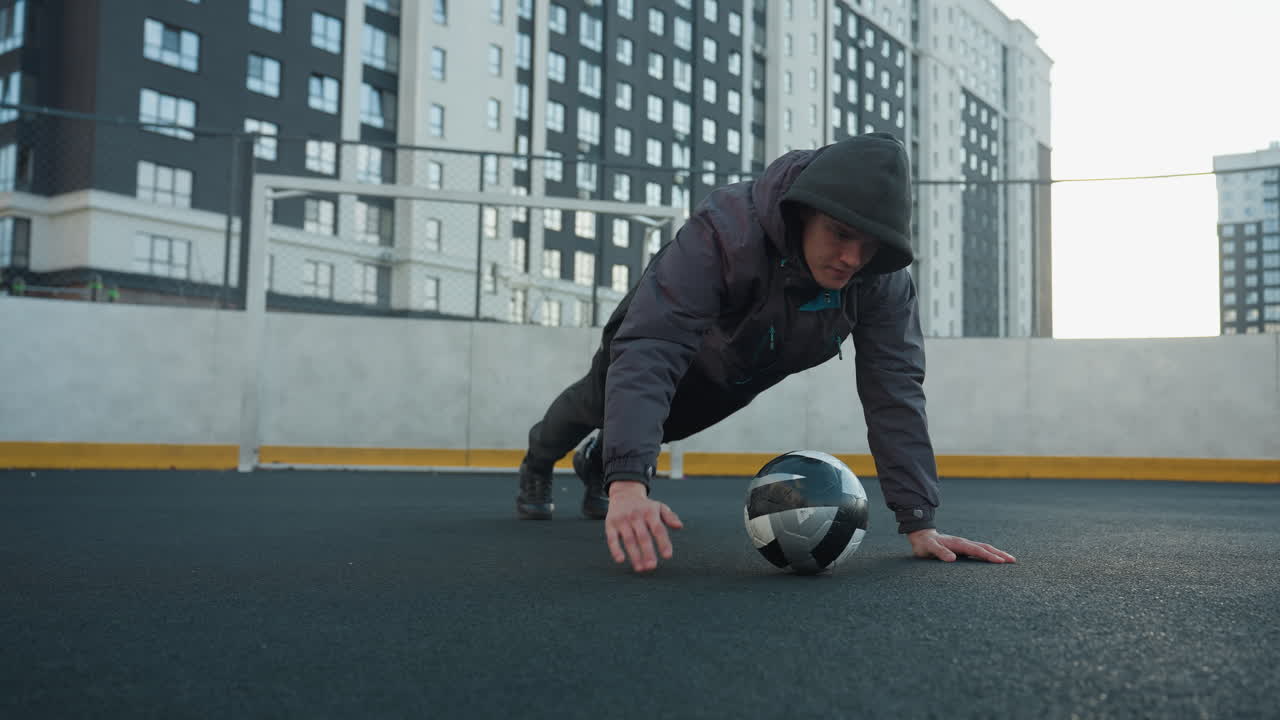 Athlete performing push-ups alternating hand placement on soccer ball demonstrating strength, coordination, and agility in outdoor urban arena with goal post and residential building background