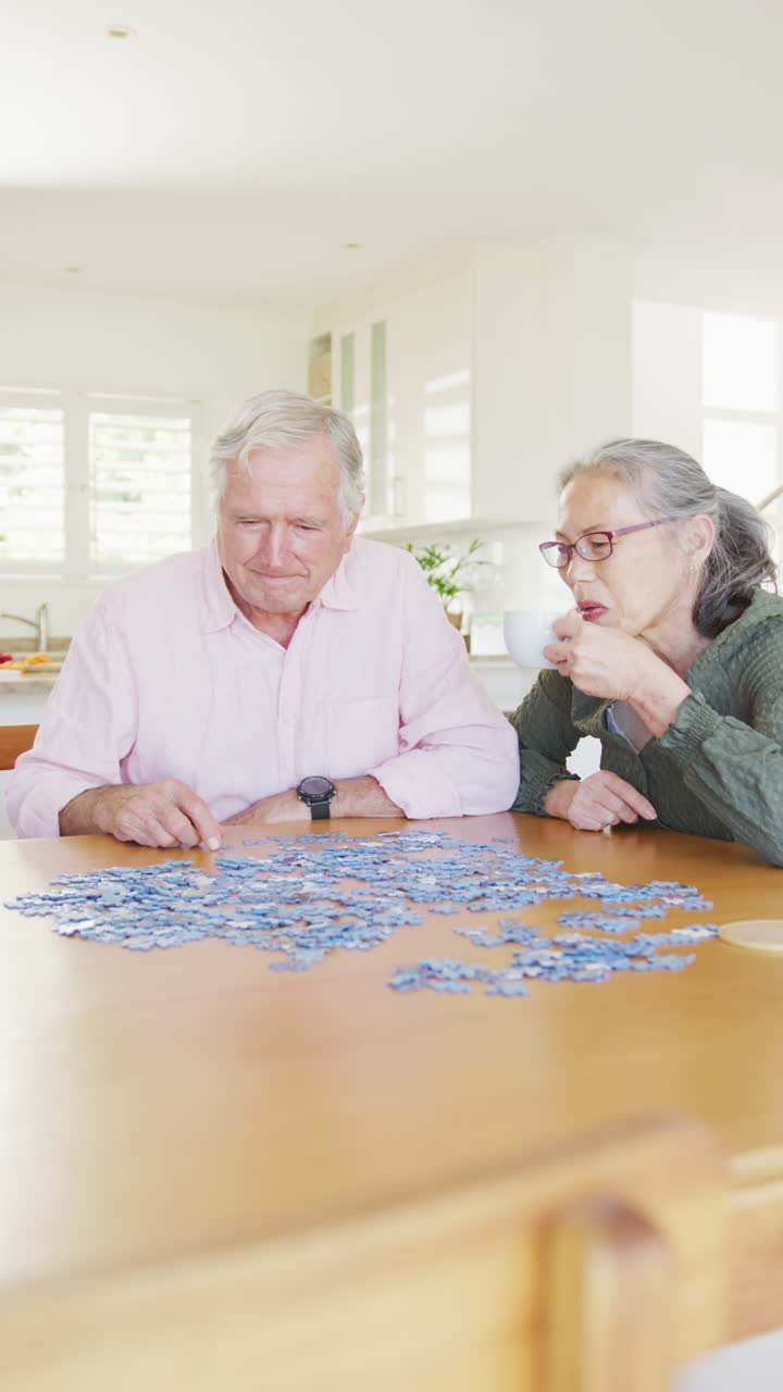 Vertical video of happy diverse senior couple sitting at table and doing puzzle