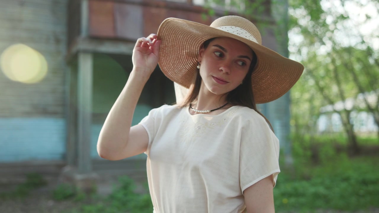 Una giovane donna si trova in un luminoso cortile-giardino indossando un cappello di paglia a tesa larga e una camicia bianca ampia, con una mano che tocca il cappello sotto la calda luce del giorno