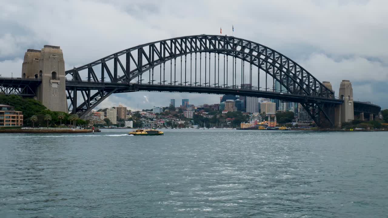 A boat passes beneath the iconic Sydney Harbour Bridge, highlighting the massive steel structure as it spans the harbor, connecting the vibrant city skyline.