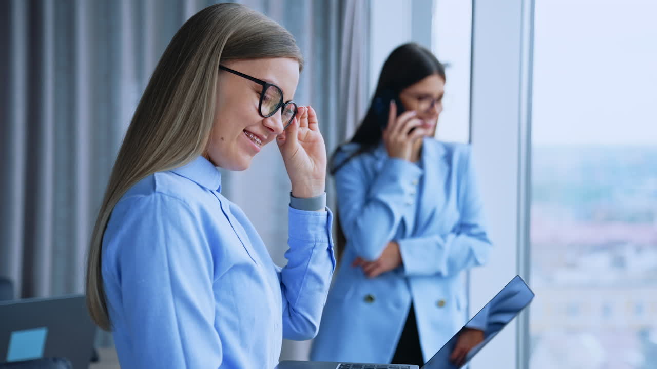 Millennial female employees using modern gadgets for communication. Blonde lady at foreground holding laptop and dark-haired lady at backdrop speaks on the phone. Blurred background.