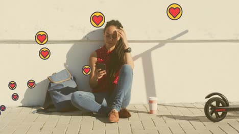 múltiples iconos de corazón flotando contra una mujer afroamericana usando un teléfono inteligente sentada en la calle