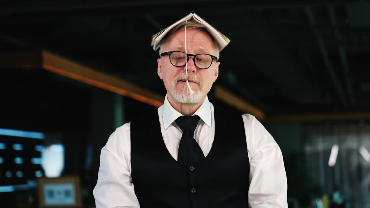 Adult Caucasian man wearing glasses holds a book on his head. Mature man fooling around in office.