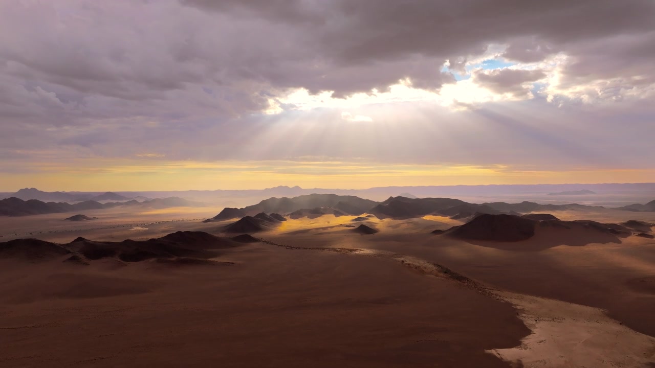 Vista drone di un drammatico cielo nuvoloso con raggi di sole sopra le dune e il bacino secco del deserto del Namib. Luce soffusa e nebbia alla deriva avvolgono le antiche dune e montagne in un'atmosfera calma e cinematografica