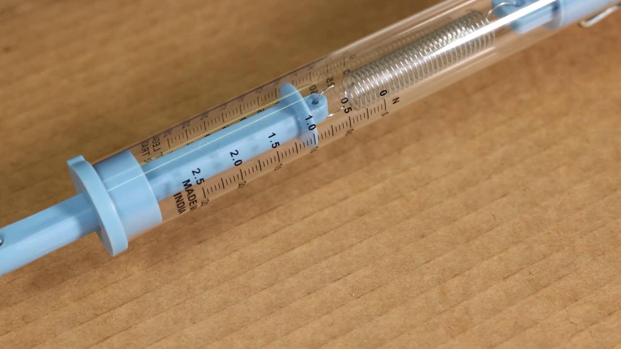 A spring scale measures friction as a wooden block is dragged across a cardboard surface, highlighting force dynamics