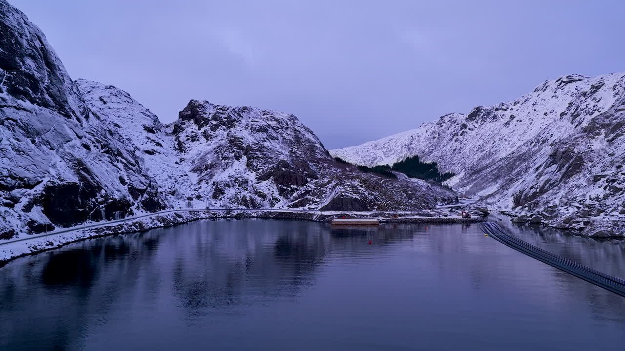 Elokuvamainen ilmakuva lumihuippuisista huipuista ja rauhallisesta vuonosta Lofooteilla, Norjassa, sinisen tunnin aikaan. Ihanteellinen matkadokumentteihin ja ympäristötarinankerrontaan.