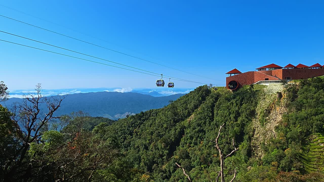 Gondole della funivia che scivolano tra le cime dei monti Ba Na Hills, oltre un edificio stazione con tetto rosso, con un'ampia valle piena di nuvole e cielo blu sullo sfondo