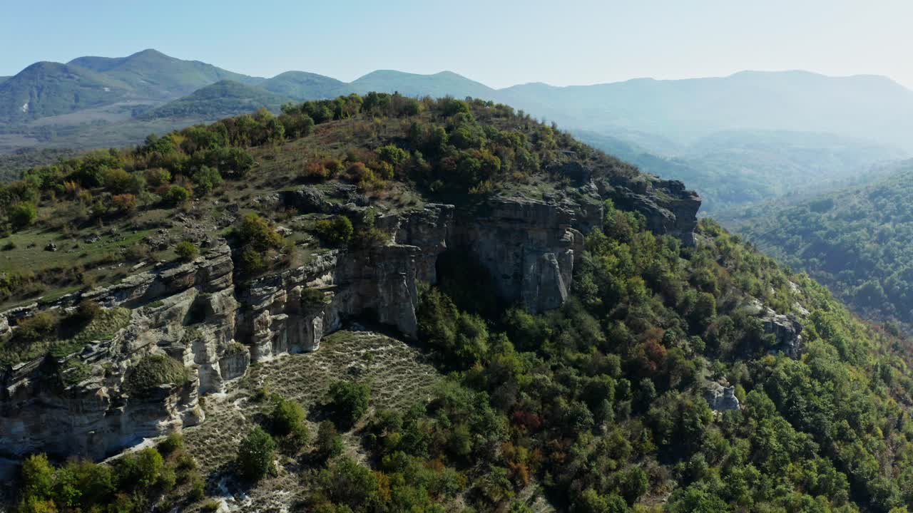 Una splendida veduta aerea di una montagna coperta da rigogliosi alberi verdi, con montagne, luce solare e cielo sullo sfondo