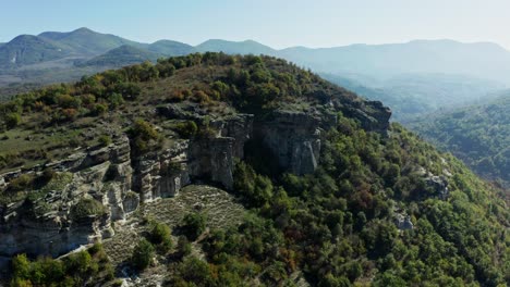 una hermosa vista aérea de una montaña cubierta de frondosos árboles verdes, con montañas, luz solar y cielo en el fondo