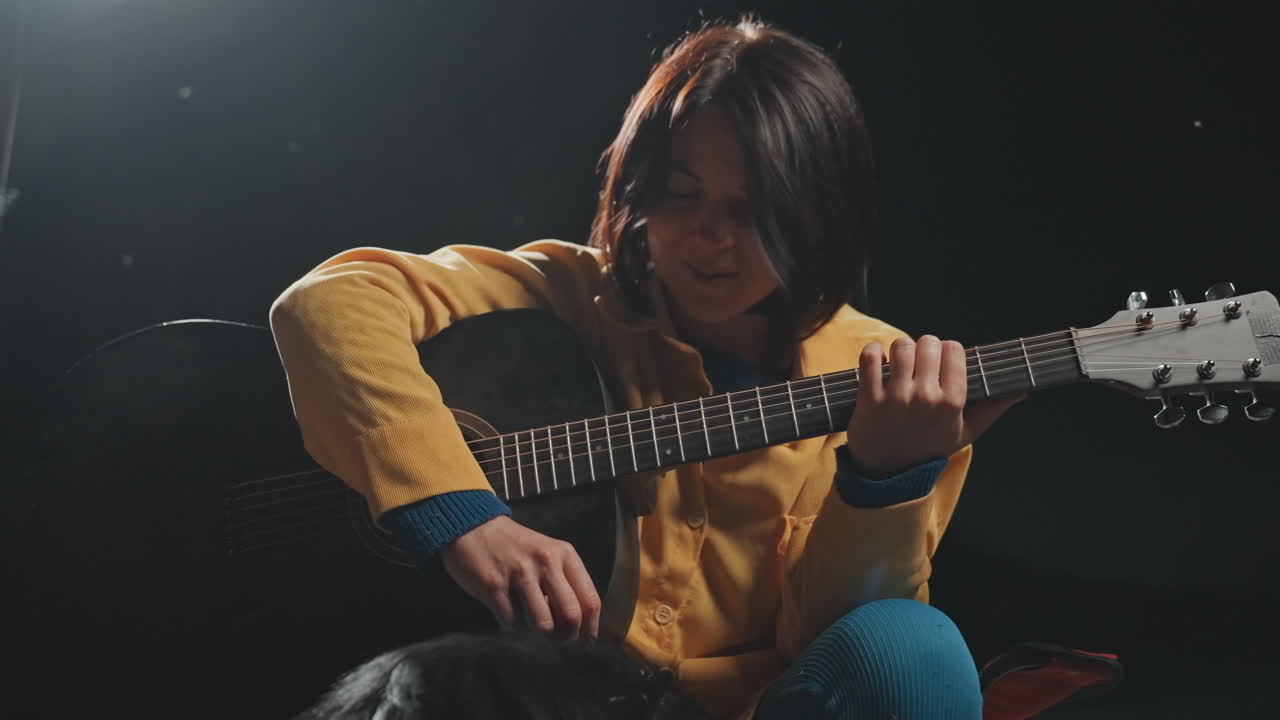 Donna in giacca gialla che suona la chitarra in campeggio, accarezza il cane sotto la luce soffusa di una lanterna, strimpellio acustico e melodia delicata, bagliore caldo di brace e luce stellare sottile, atmosfera intima e rilassata, atmosfera rustica all'aperto.
