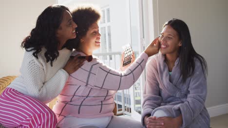 Happy-diverse-female-friends-doing-make-up-and-smiling-in-bedroom