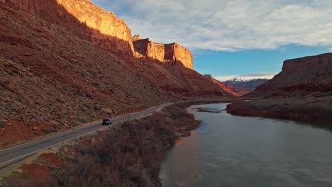 wide rearview tracking of car driving below of layered red rock cliffs with desert floor and deep blue sky overhead in clear light, moab utah usa