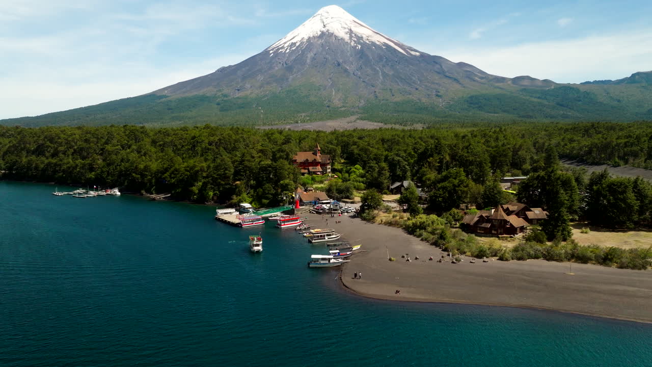Forward-moving drone glides across Lake Todos los Santos toward a small harbor, as snowcapped Osorno volcano rises beyond forest and dark sand shore beneath a clear summer sky