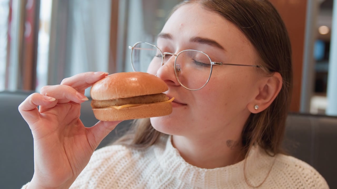 Young woman lifts cheeseburger to mouth and takes bite in sunlit cafe booth, eyes closed and slight smile expressing enjoyment, while raised hand, soft posture, and warm interior set casual dining