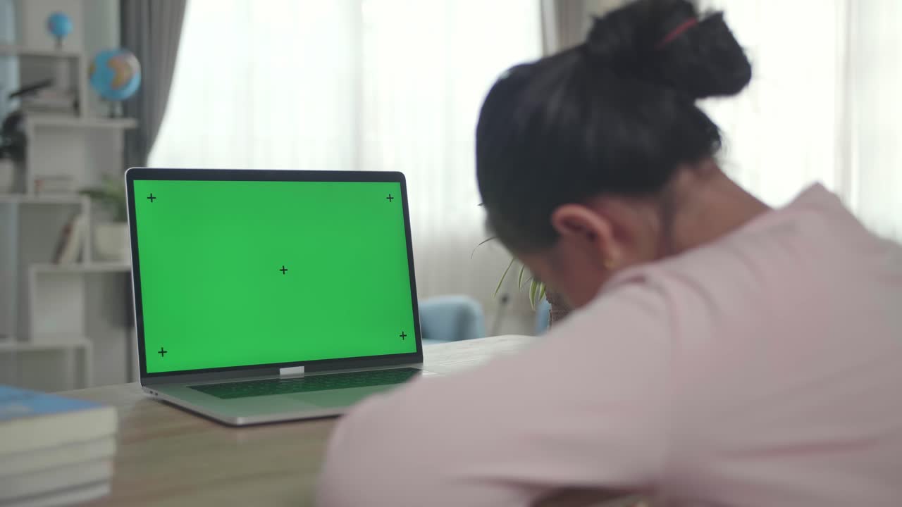 Teenage Girl Sitting At Her Desk Learning Online On A Laptop With Green Mock-Up. Over Shoulder Close Up View