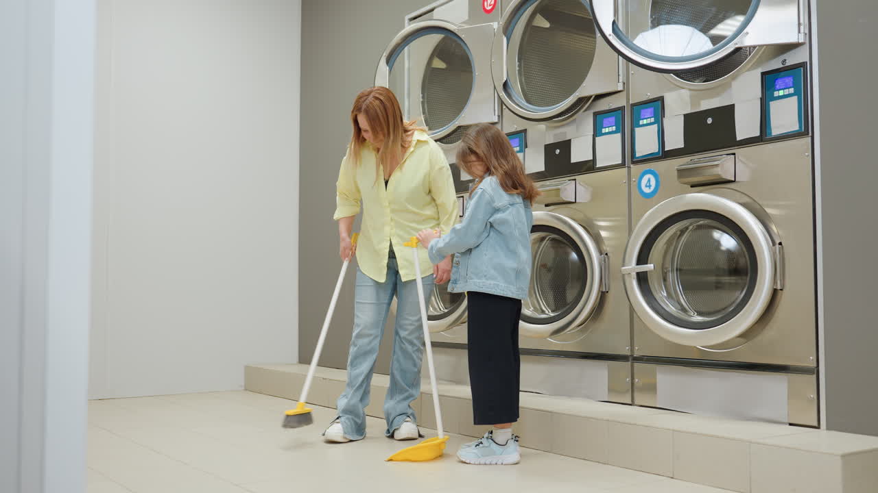 Youngster holds dustpan while parent sweeps tiled floor in laundromat near stainless industrial washers, family cleaning showing guidance, responsibility, hygiene, routine care during chore scene