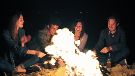 Group-of-young-multiethnic-people-roasting-marshmallow-on-sticks-over-the-fire-together-on-the-beach-late-at-night,-drinking-beer