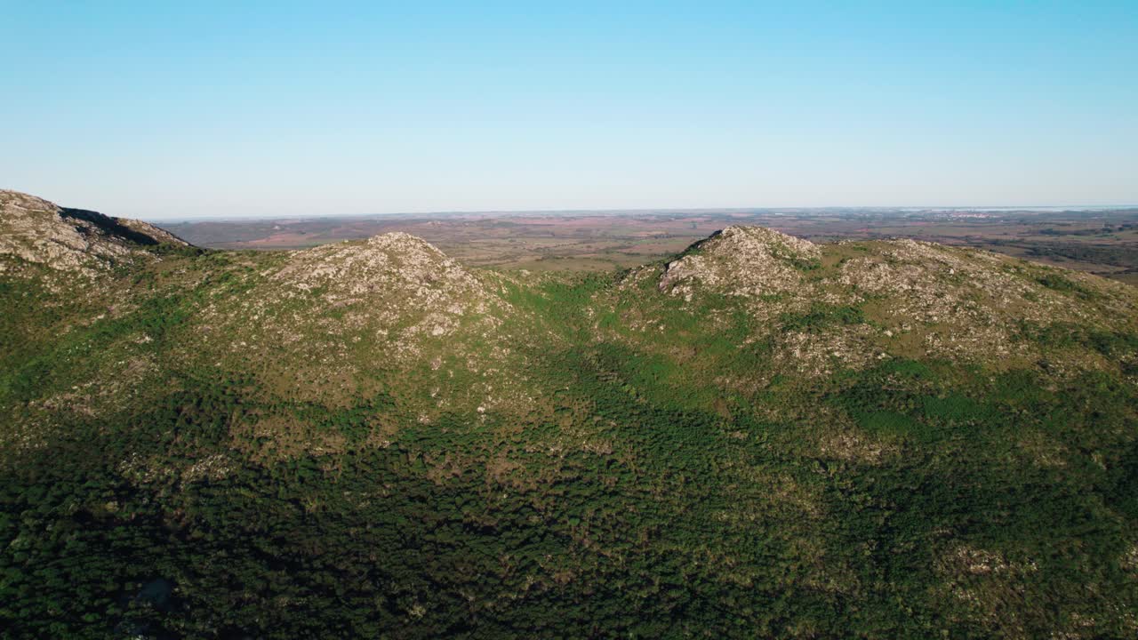 An aerial drone shot capturing the rocky, exposed granite hills and the dense forest cover of the Uruguayan countryside. Features a clear blue sky and vast, natural landscape