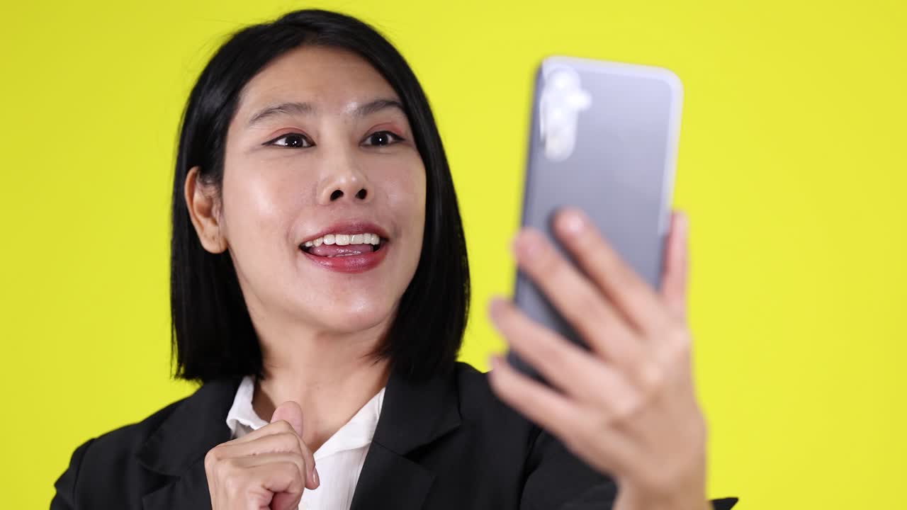 Woman in black blazer expresses surprise, excitement, and ideas on smartphone video call, yellow background