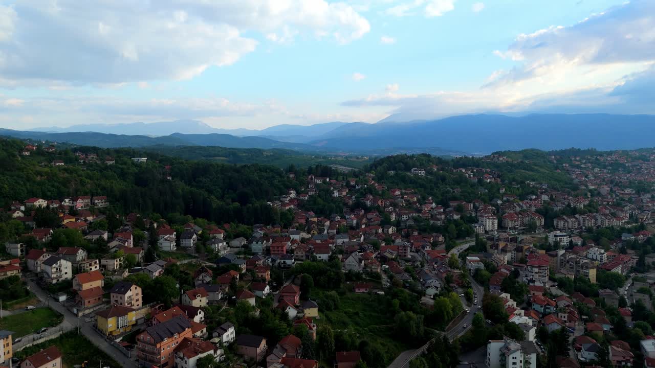 Góc nhìn từ trên không bằng máy bay không người lái về vùng ngoại ô Sarajevo ở Bosnia và Herzegovina, cho thấy những ngôi nhà mái đỏ trải rộng trên những ngọn đồi xanh với những ngọn núi ở đằng xa dưới ánh sáng dịu nhẹ buổi chiều