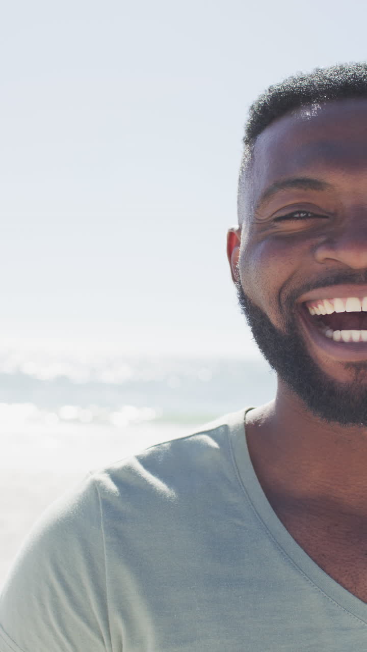 Vertical video of happy african american man on beach smiling at camera