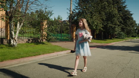 little girl in blue gown with long hair walking alone along quiet residential street, holding soft toy and glancing at tall iron fence on right side as sunlight casts clear shadow on ground