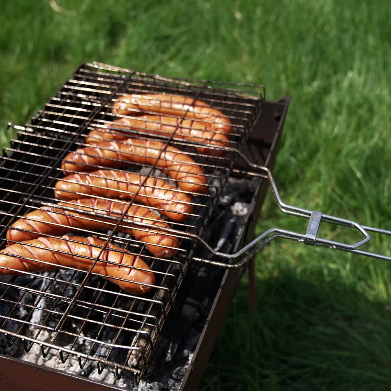 Roasting sausages on a grill outdoors. Delicious hot dog sausages grilling in smoke among green nature. Cooking meat sausages for a picnic