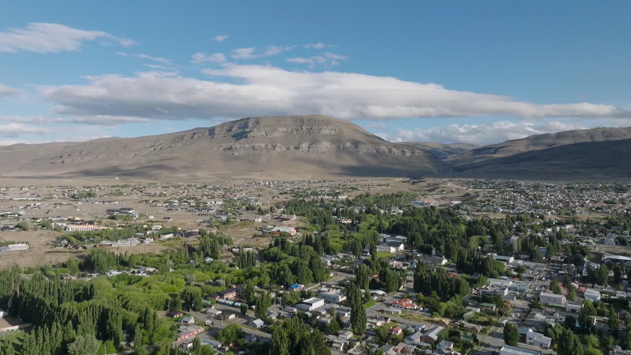 Aerial drone footage of Calafate town nestled between desert mountains and lush green residential areas. Sweeping forward movement showcases dramatic Patagonian landscape.