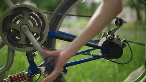 close-up partial view of someone s hand rotating a bicycle's wheel, focusing on the chain and gear system, the background features a softly blurred view of greenery