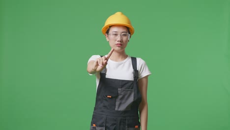 asian woman worker wearing goggles and safety helmet pointing at you and disapproving with no index finger sign while standing in the green screen background studio