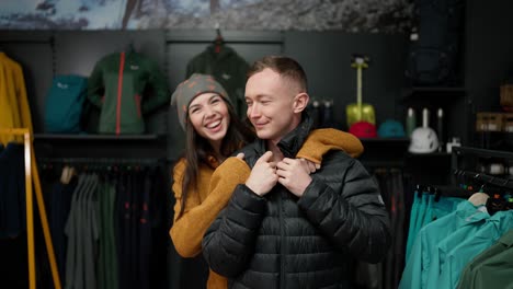 young man trying at winter jacket in clothing store with his girlfriend, front view