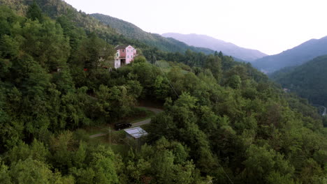 Schöne-Drohnenaufnahme-Aus-Der-Luft-Eines-Großen-Hauses,-Das-Mitten-In-Einem-Wald-Auf-Einem-Berg-Steht,-Schwarzes-Auto-Fährt-Eine-Schmale-Straße-Hinauf-Zum-Haus,-Drohne-Fliegt-Langsam-Näher