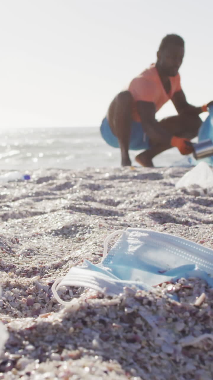 아프리카계 미국인 남자가  ⁇ 은 해변에서 장갑으로 쓰레기를 분리하고 있습니다.