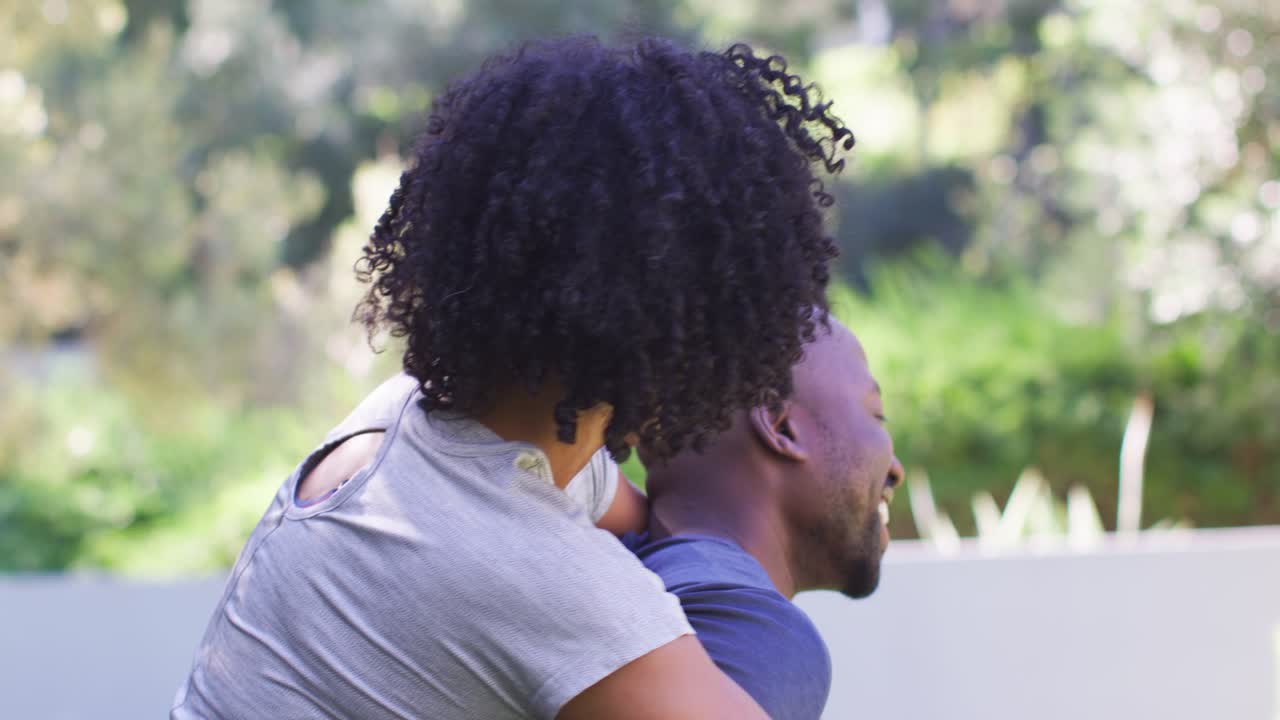 African American couple playfully laughing, piggybacking in yard by low wall, woman leaning on man