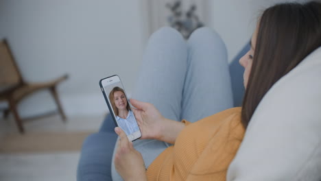 Curly-young-woman-having-video-chat-with-friends-using-mobile-phone-camera-while-lying-on-bed.-Attractive-Cheerful-Woman-Sitting-On-The-Couch-And-Having-A-Video-Call-With-GirlFriends