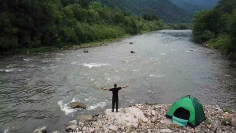 the man demonstrates a sense of freedom arms outstretched the man demonstrates a sense of freedom arms outstretched resting in the mountains near a stormy river green tent